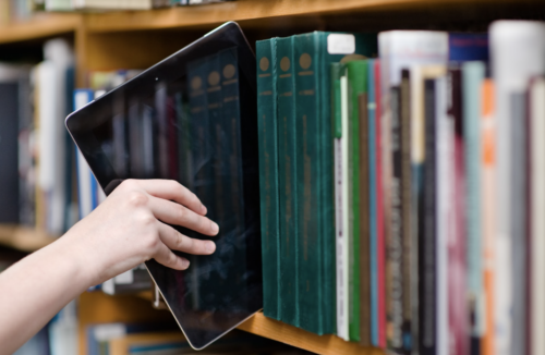 a hand pulling a tablet off a library shelf filled with books