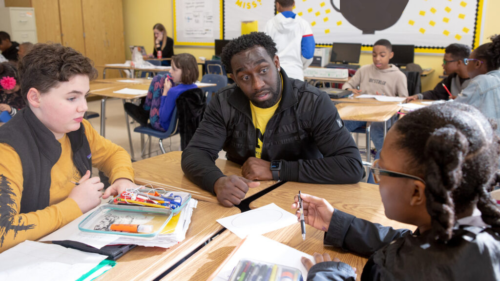 A teacher sits at a classroom table, talking with two middle school students